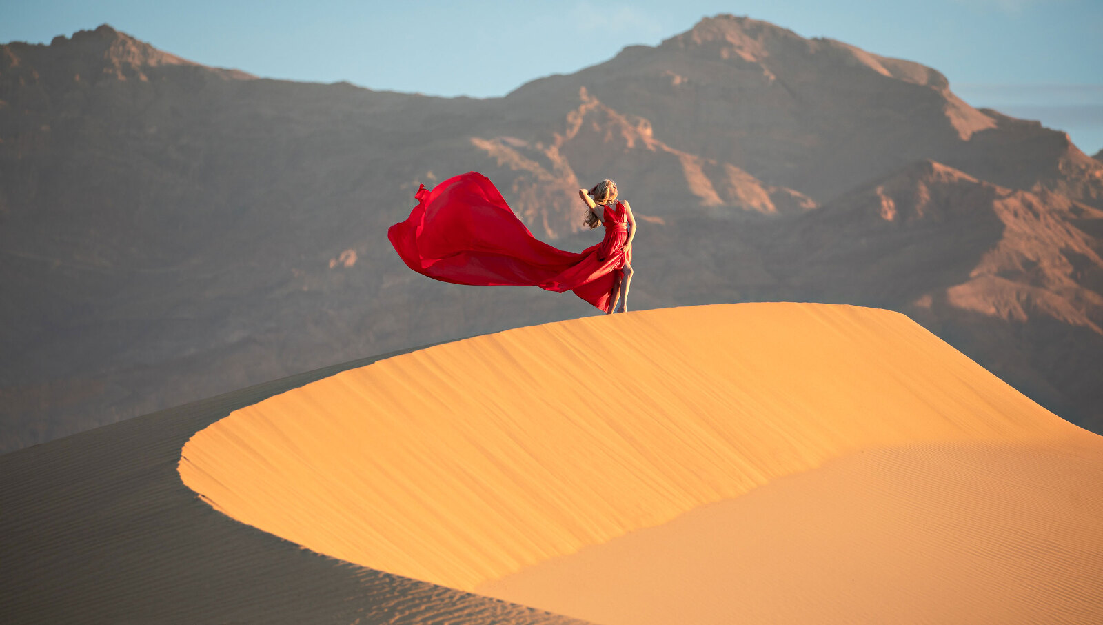 Woman on top of sand dune in a red, long dress with mountain.