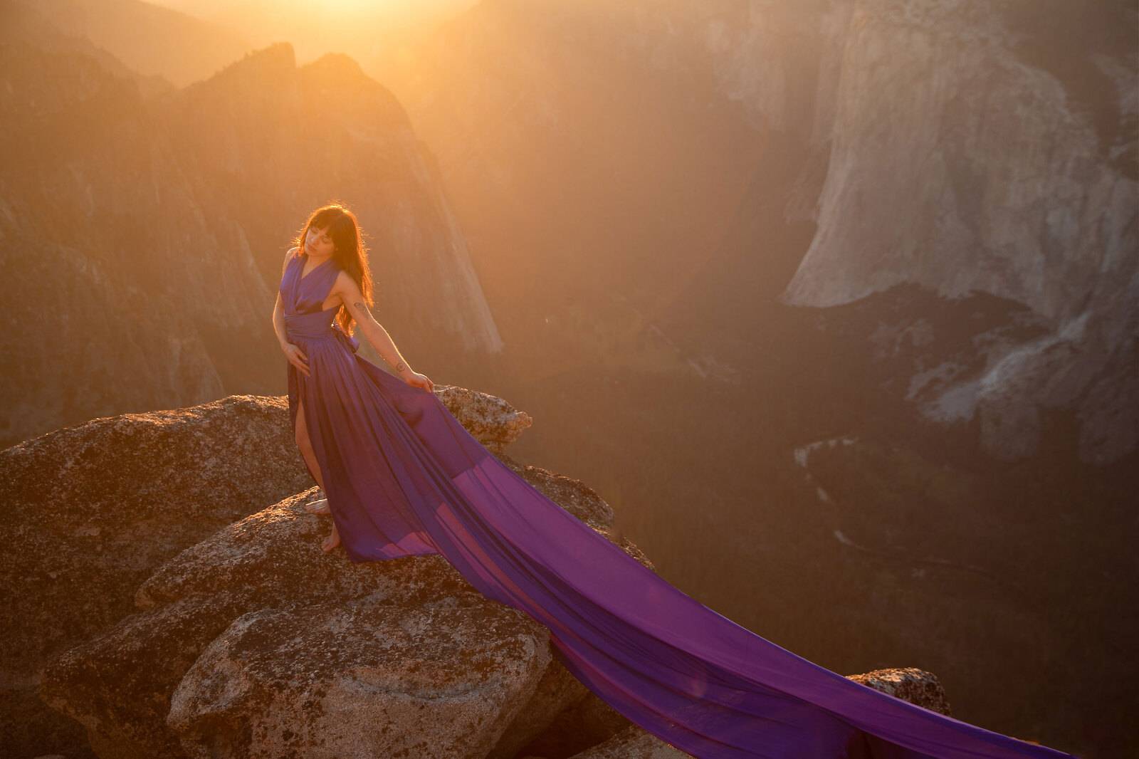 Woman at a viewpoint in a long, purple dress during sunset with mountains.