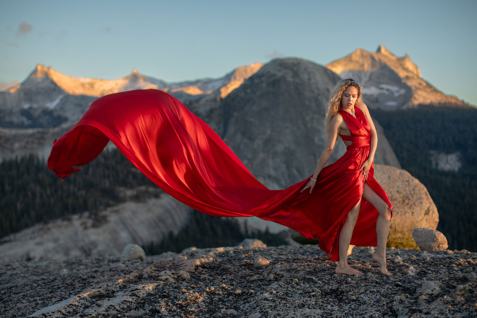 A woman in a long, red dress stands on top of dome at sunset with mountains.