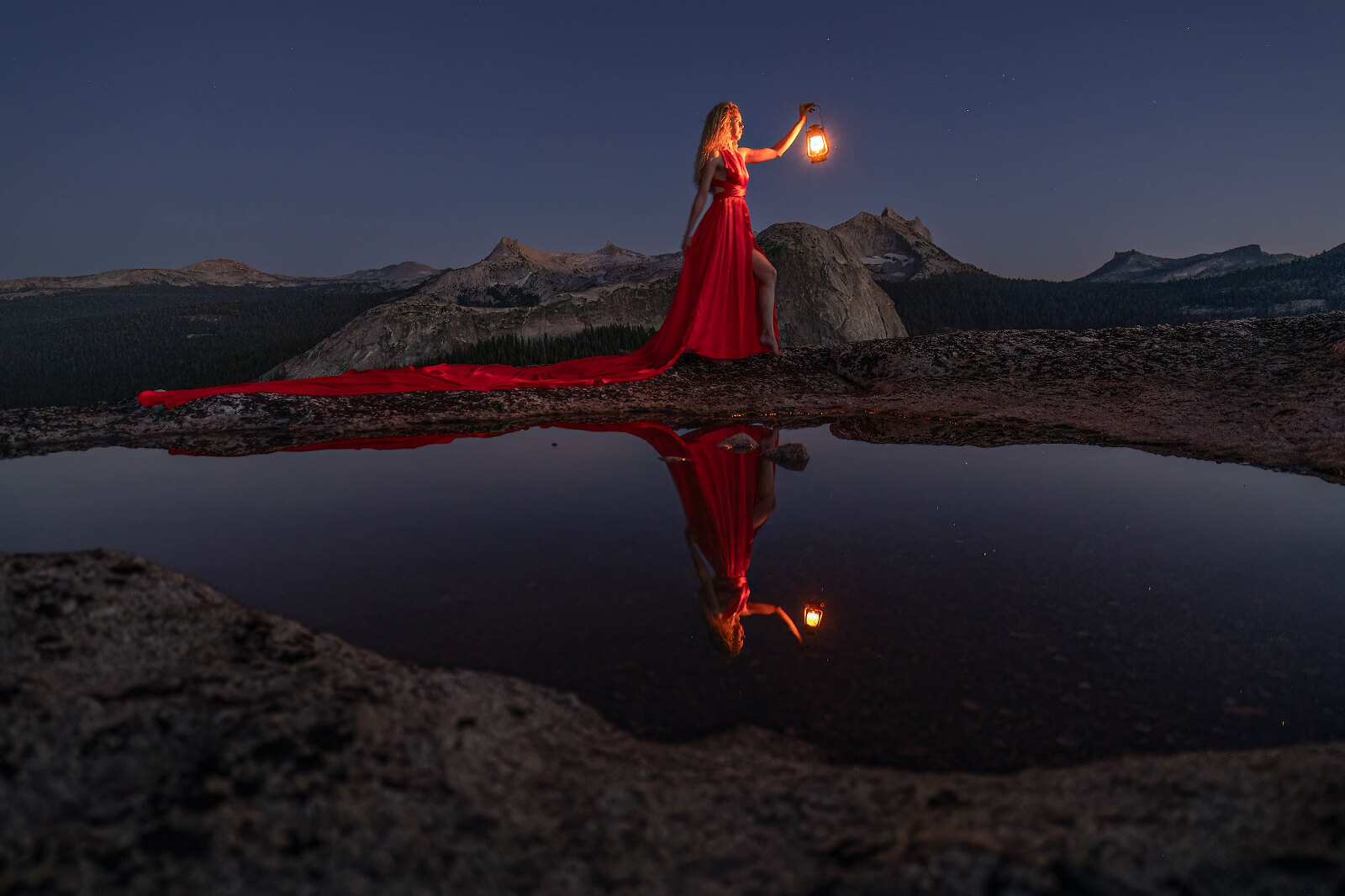 A woman wearing a long, red dress stands next to a pool of water with mountains at night.