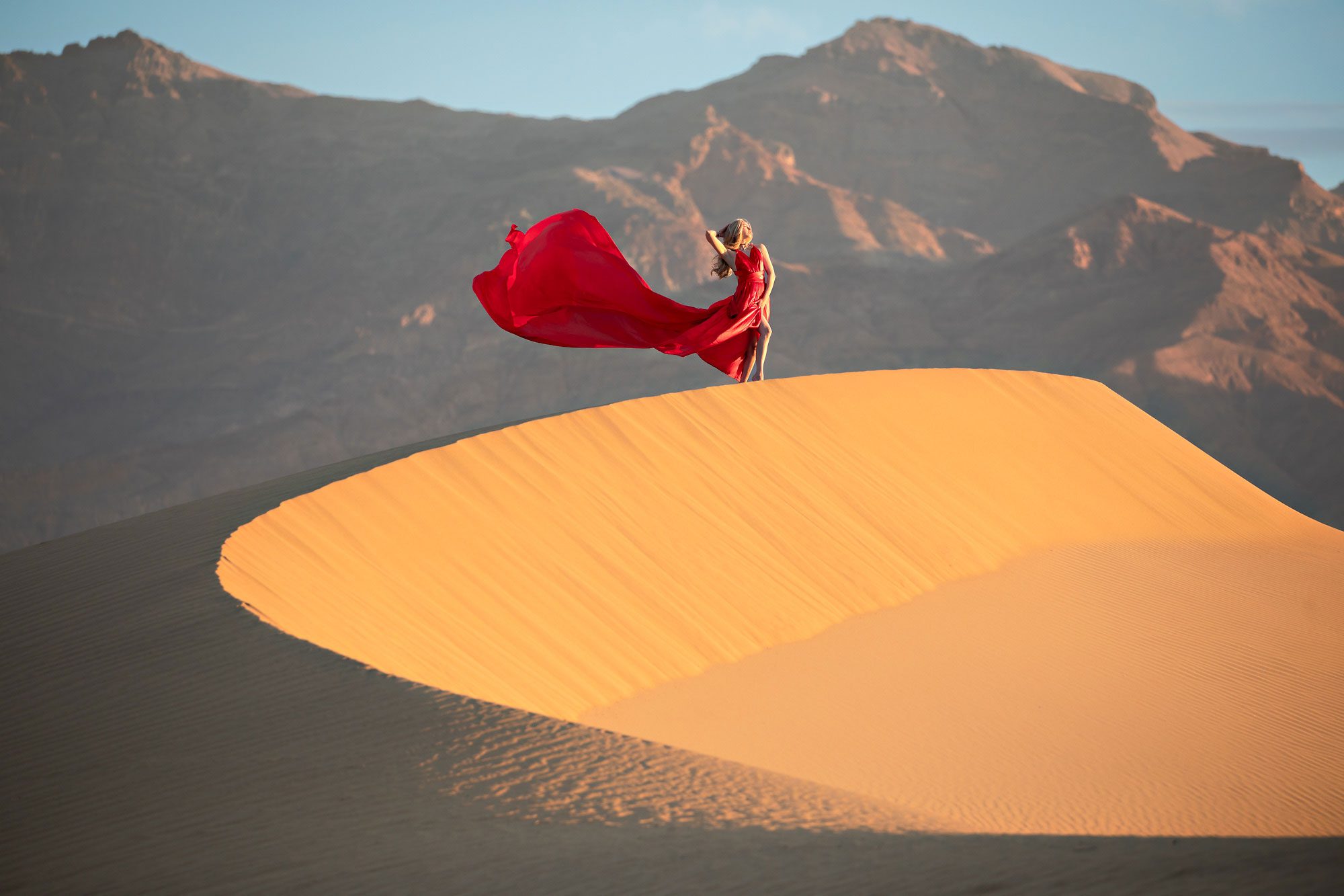 Woman wearing a red dress standing on top of sand dune with mountains and blue sky.