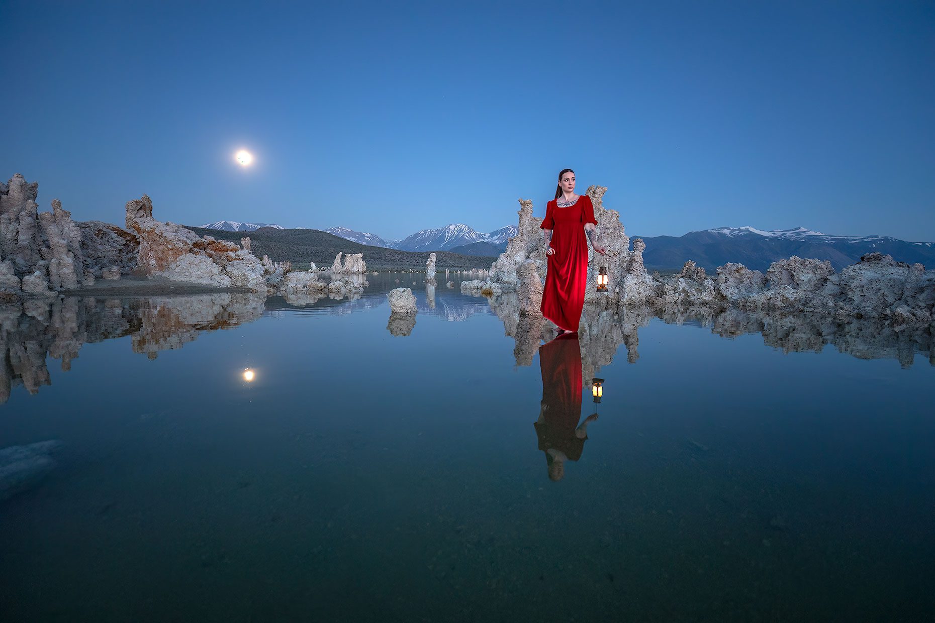 A woman standing in water with a reflection at dusk with the moon and mountains.